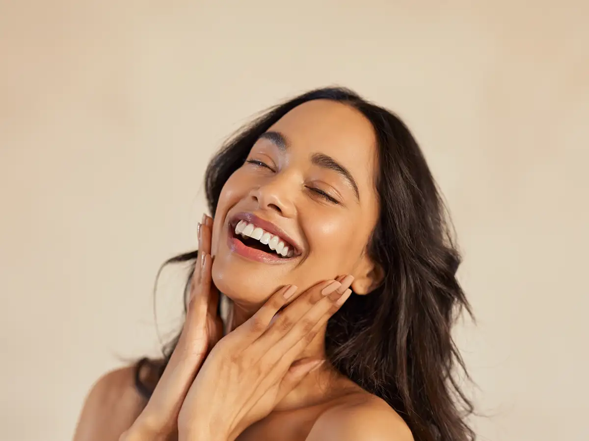 A smiling woman with radiant, smooth skin gently touching her face, showcasing the natural, non-frozen results of a wrinkle relaxer treatment.
