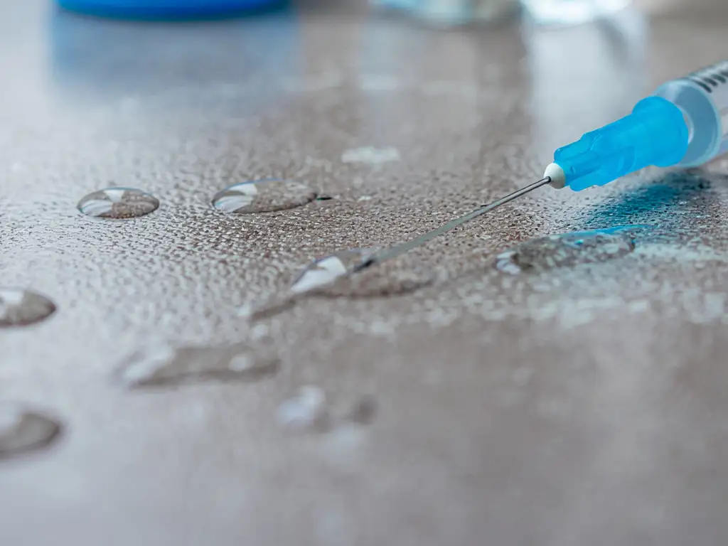 Close-up of a medical syringe and needle resting on a textured surface surrounded by clear droplets of liquid silicone.